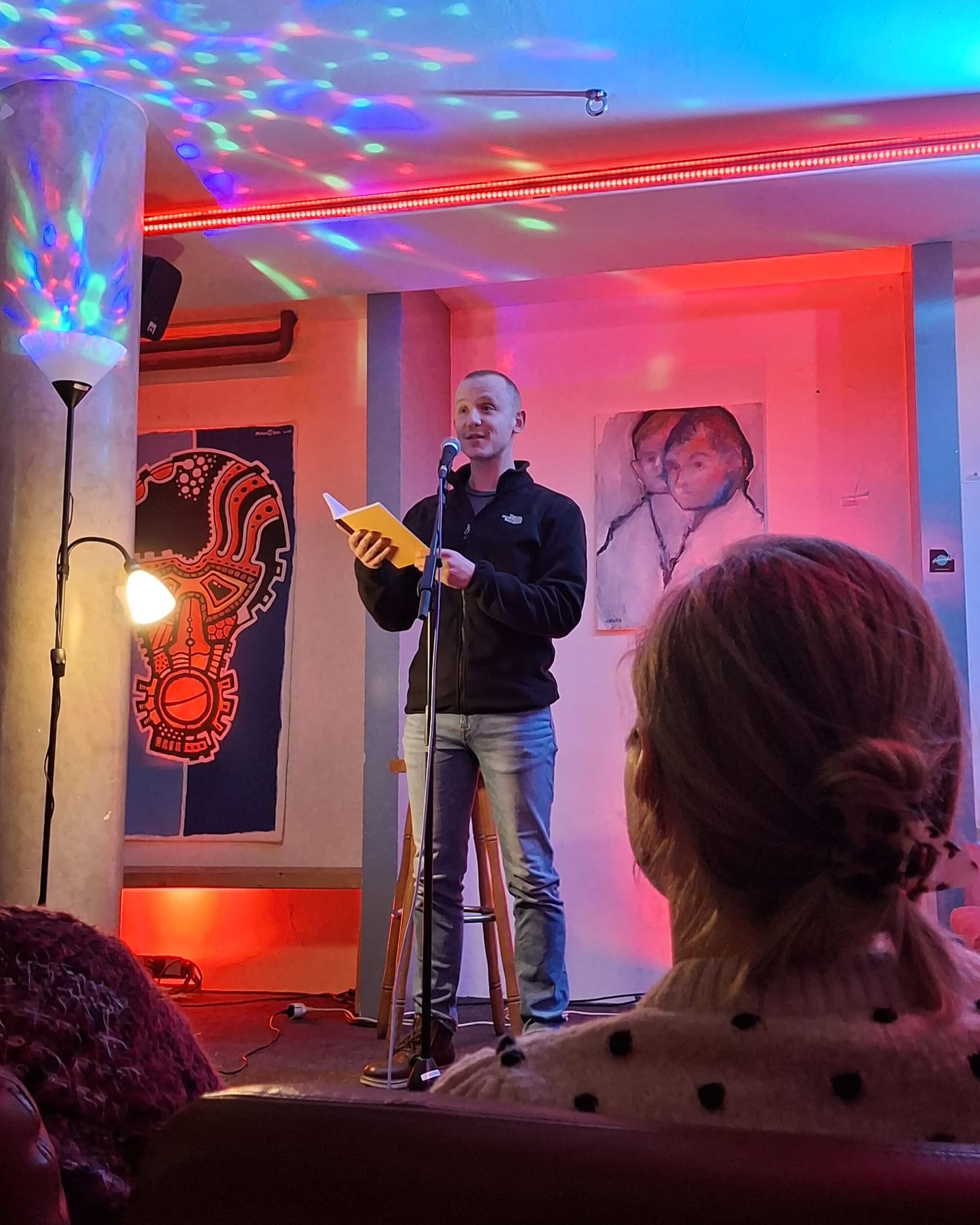 White man with blonde hair standing on a stage reading from a paper notebook into a microphone. People listening.